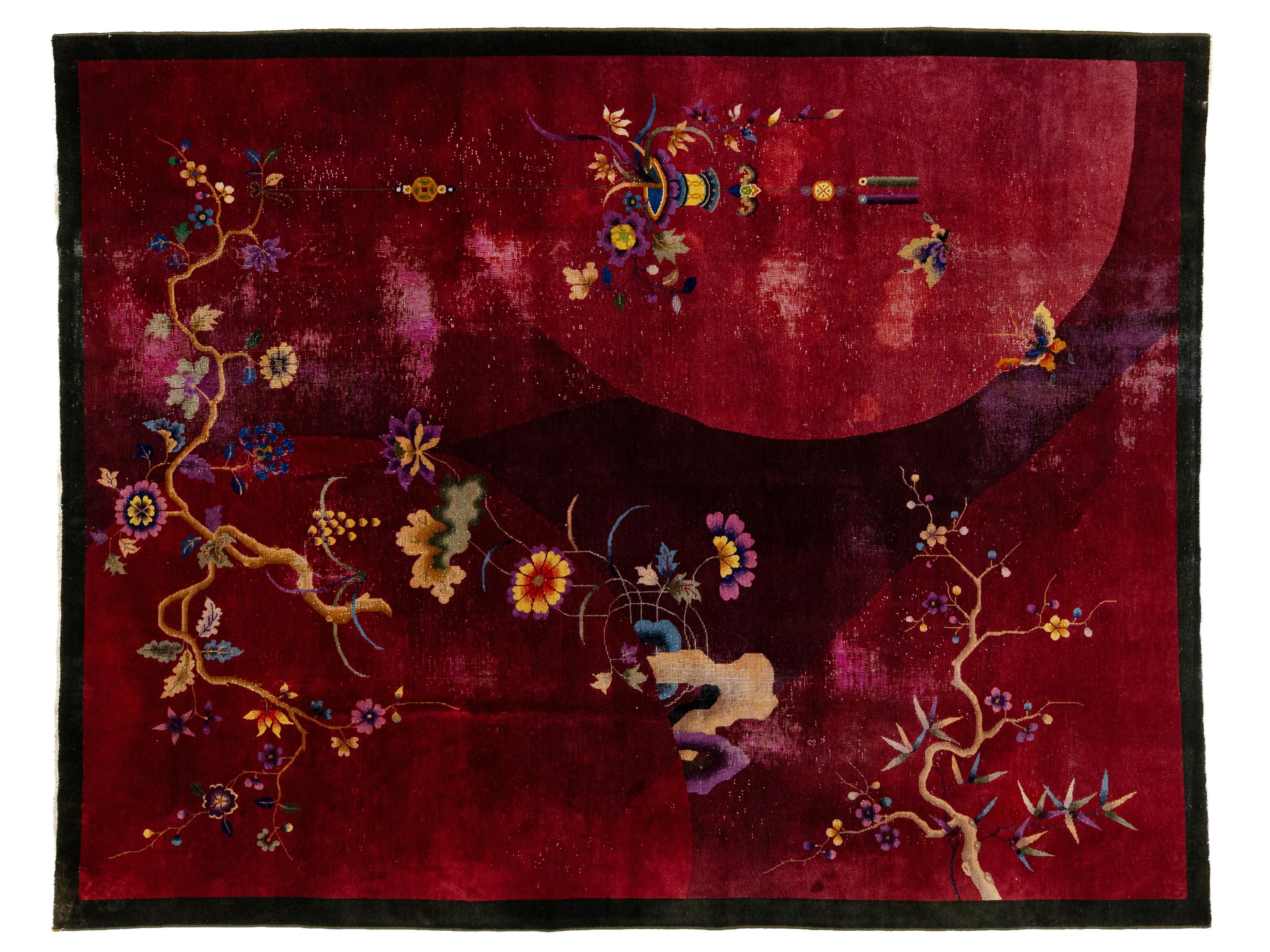A square red textile or rug with a black border and embroidered floral motifs in gold, blue, purple, and yellow, plus a large pale red circular shape in the upper right.