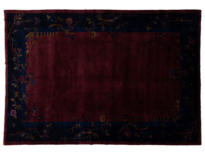 A rectangular area Chinese rug with a deep burgundy center field and a dark navy blue border decorated with floral and vine motifs in muted gold, pink, and blue tones.