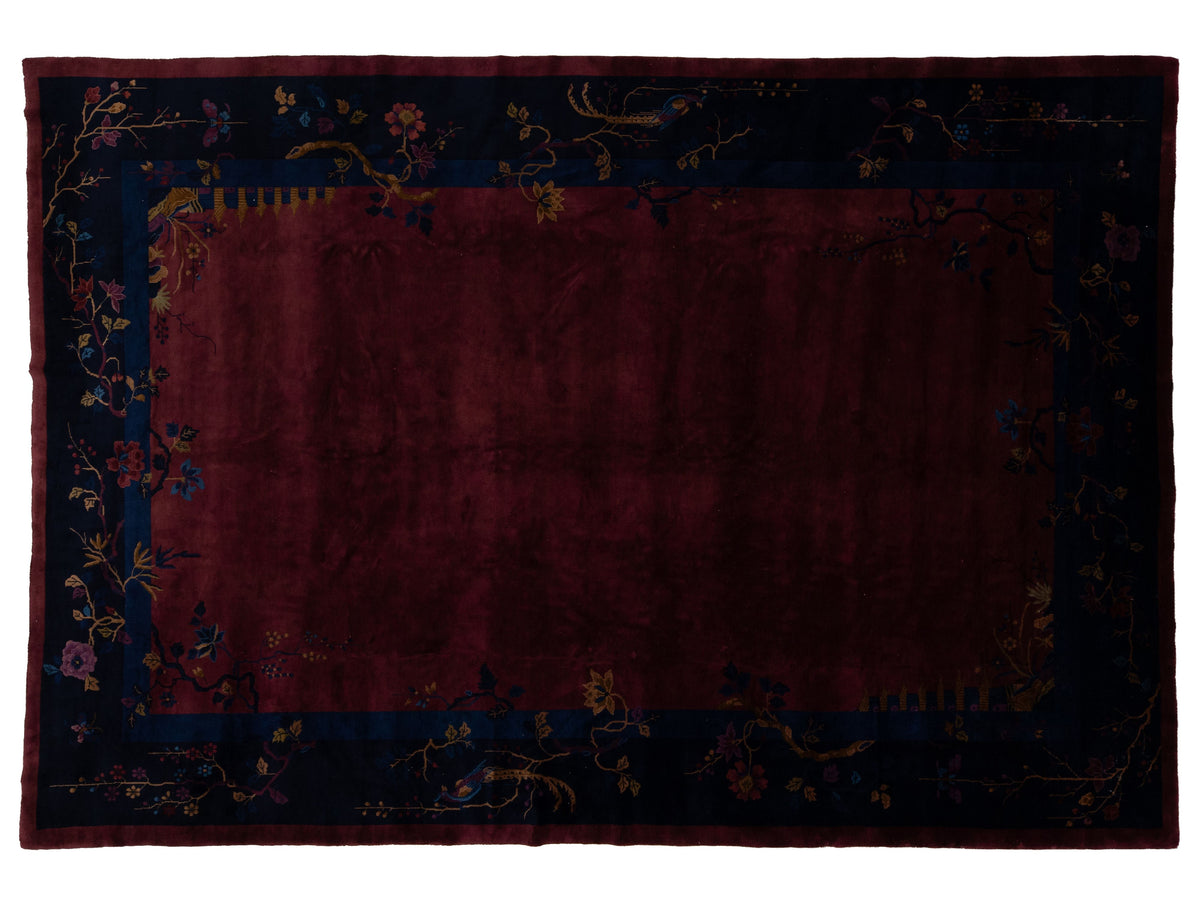 A rectangular area Chinese rug with a deep burgundy center field and a dark navy blue border decorated with floral and vine motifs in muted gold, pink, and blue tones.