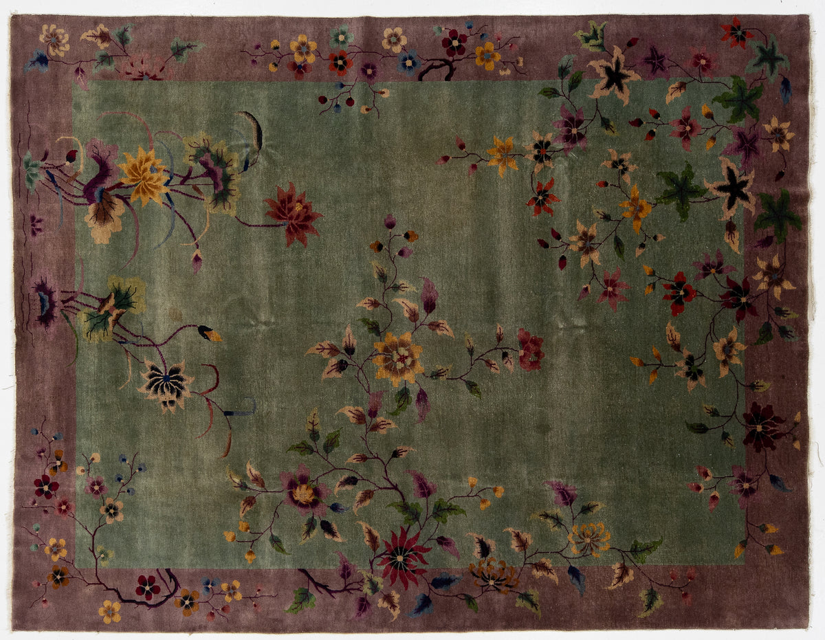 A rectangular Chinese floral rug with a muted olive green center field, a dusty mauve-purple border, and scattered embroidered or woven floral motifs in red, yellow, blue, and green.