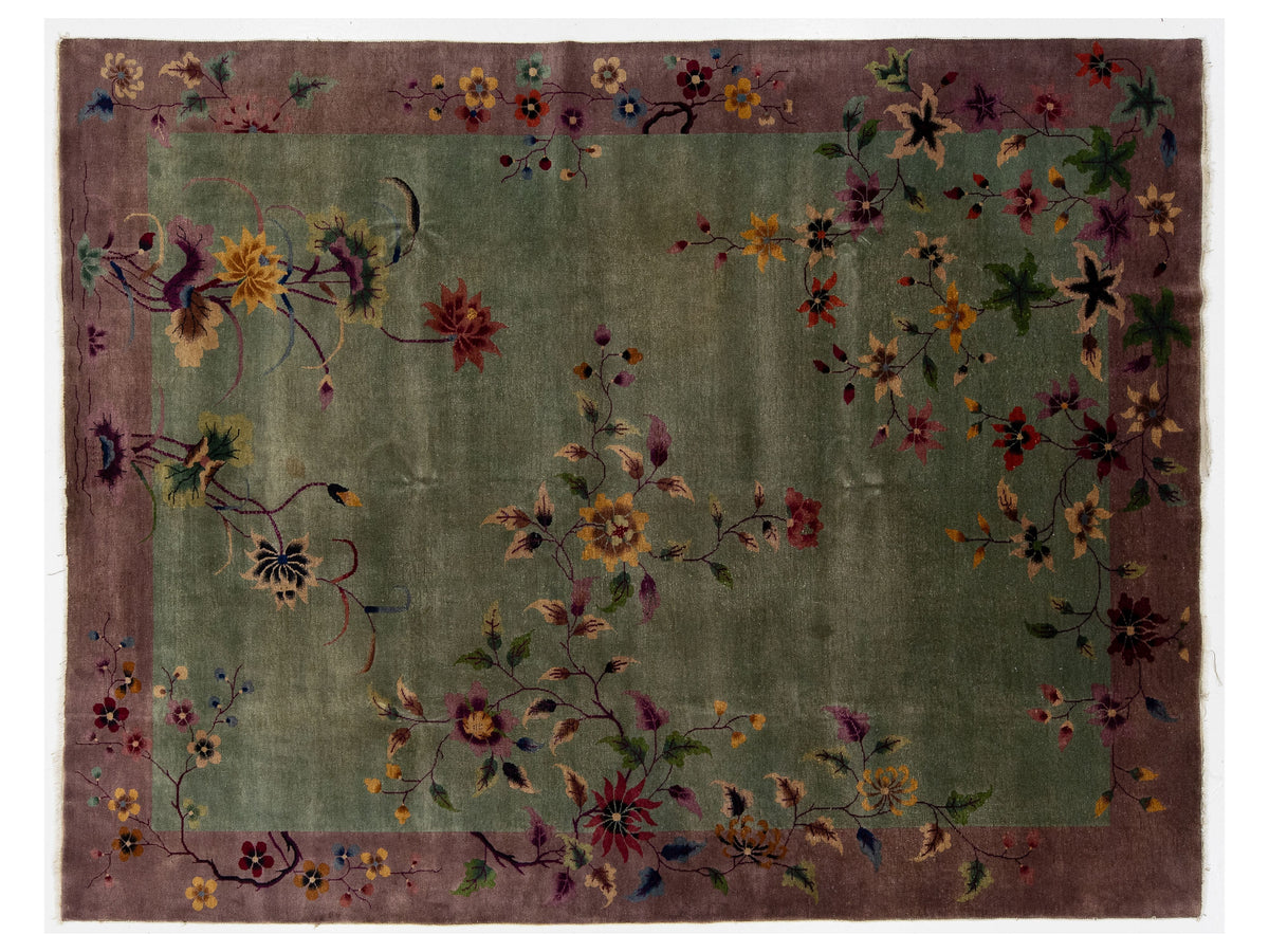 A rectangular Chinese floral rug with a muted olive green center field, a dusty mauve-purple border, and scattered embroidered or woven floral motifs in red, yellow, blue, and green.