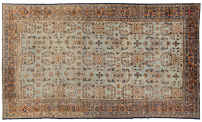 A rectangular traditional area rug with an ornate central field and detailed border, featuring a muted light blue-gray base with rust orange, beige, and navy blue patterns.