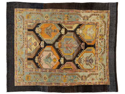 A rectangular vintage-style rug with a complex geometric and tribal pattern, featuring a central field of orange, beige, and blue motifs surrounded by a light blue inner border and a dark charcoal outer border.