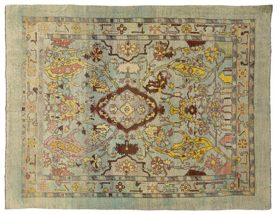 A square vintage area rug with an intricate floral and medallion pattern, featuring a muted sage green background with yellow, brown, pink, and blue accents.