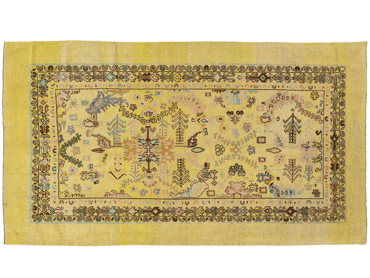 A rectangular hand-knotted wool rug with a yellow border and a central field featuring a multicolor folk-art pattern of trees, animals, and geometric motifs.