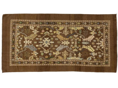 A rectangular brown area rug with an intricate, multi-colored traditional pattern featuring floral motifs, geometric borders, and stylized animal and tree designs.