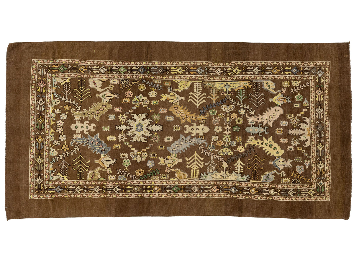 A rectangular brown area rug with an intricate, multi-colored traditional pattern featuring floral motifs, geometric borders, and stylized animal and tree designs.