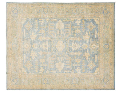 An oushak-style rectangular area rug with a traditional pattern, featuring a dusty blue central field and a warm beige border.